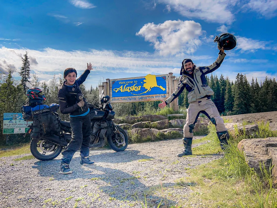 Nay e Ton comemorando ao lado da moto, em frente à placa de boas-vindas do Alasca